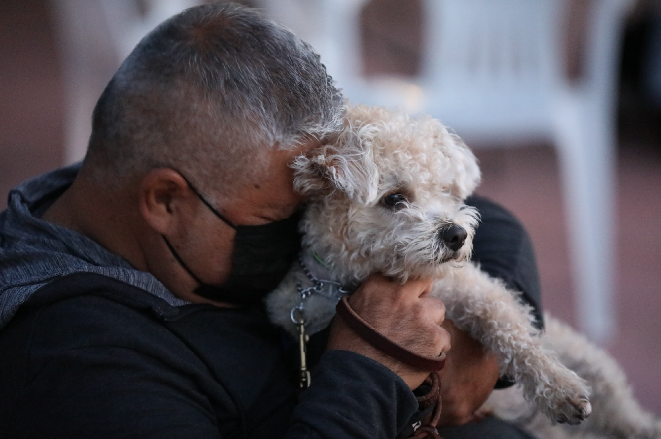 Man wearing mask holding small white dog and hugging it with his eyes closed.